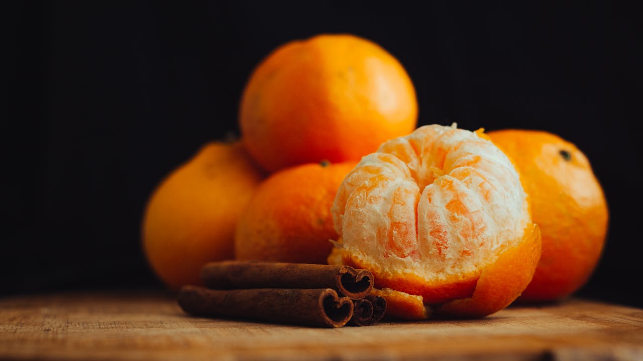 Peeled orange next to whole oranges and cinnamon sticks on a wooden surface
