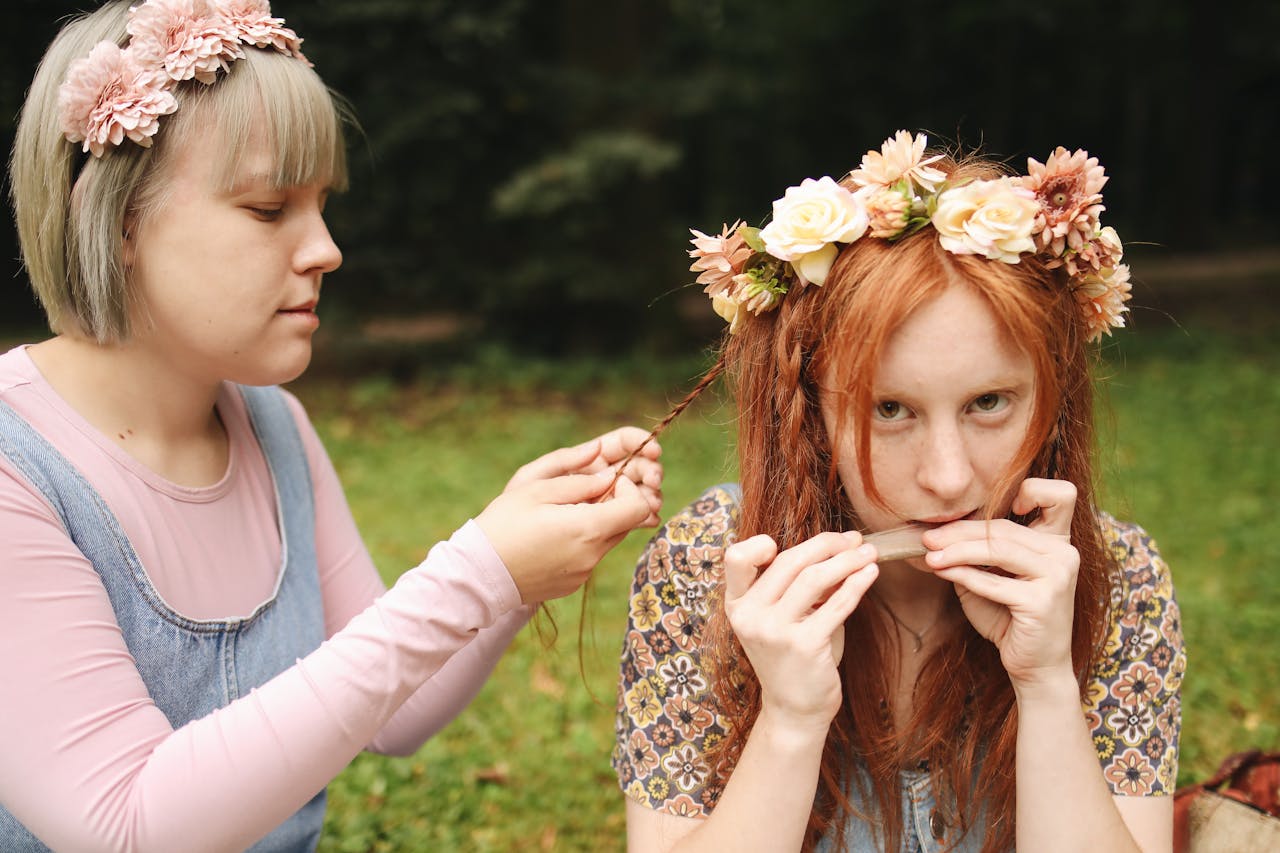 Two women outdoors; one braiding the other’s hair, both wearing dried flower crowns