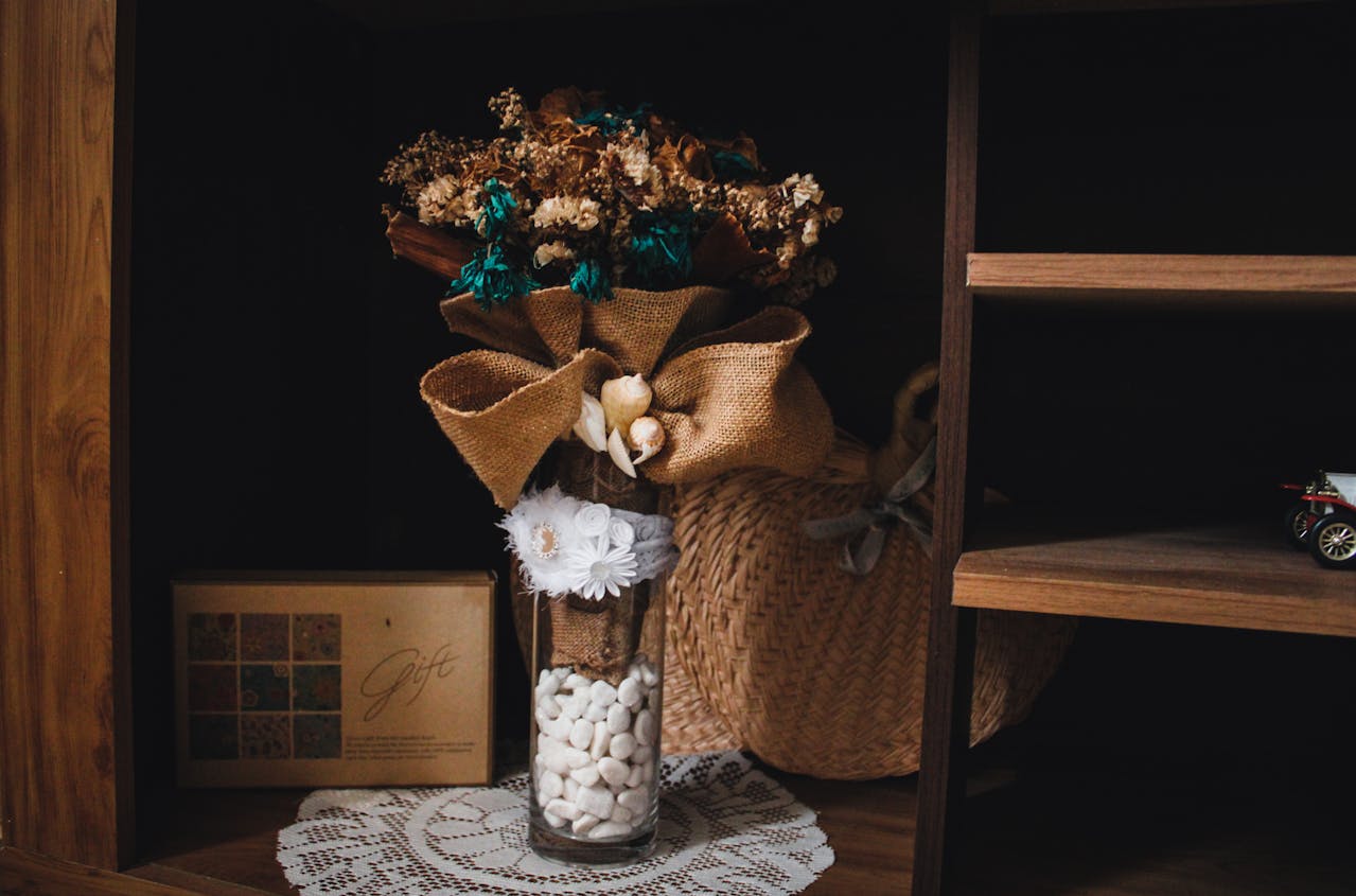 Glass vase with dried flowers, burlap bow, white stones, and lace trim on a wooden shelf