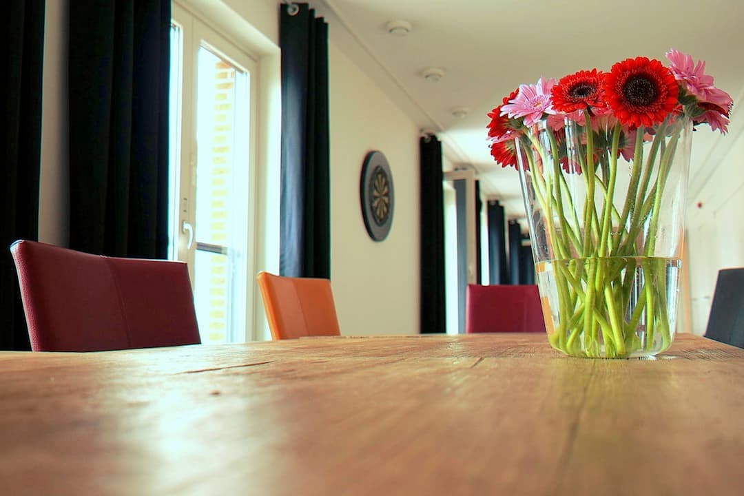 glass vase with red and pink gerbera daisies on wooden table, colorful dining chairs, long room with large windows and black curtains, wall clock in background