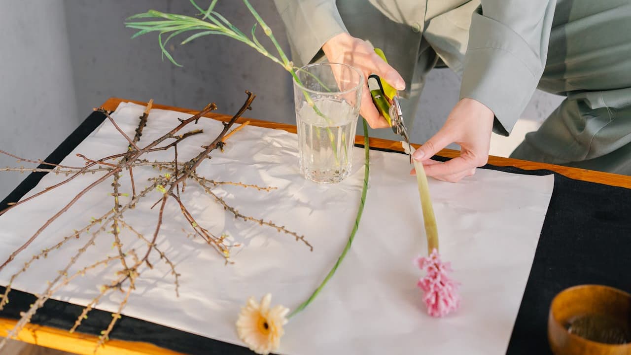 person in green outfit cutting flower stem with garden shears, glass of water and various flowers and branches on table with white paper