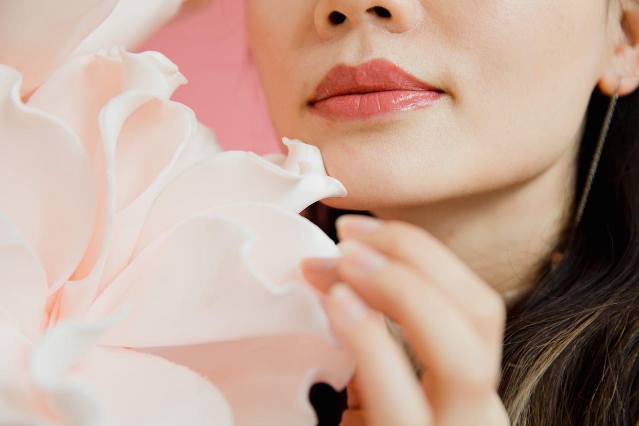 woman's lower face, glossy pink lips, hand touching large soft white flower, smooth skin, subtle makeup, pink background