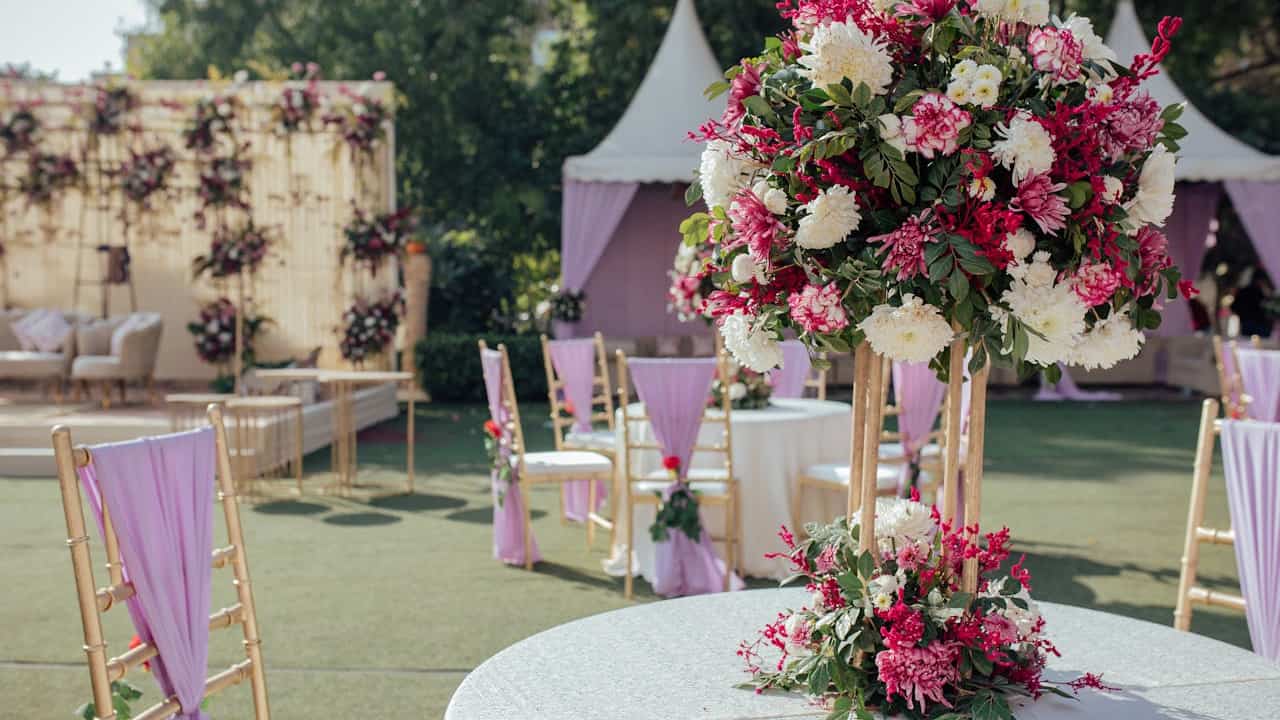 outdoor wedding setup, round tables with large floral centerpieces of white and pink flowers, gold chairs draped with lavender fabric, white tents in background, stage with floral backdrop