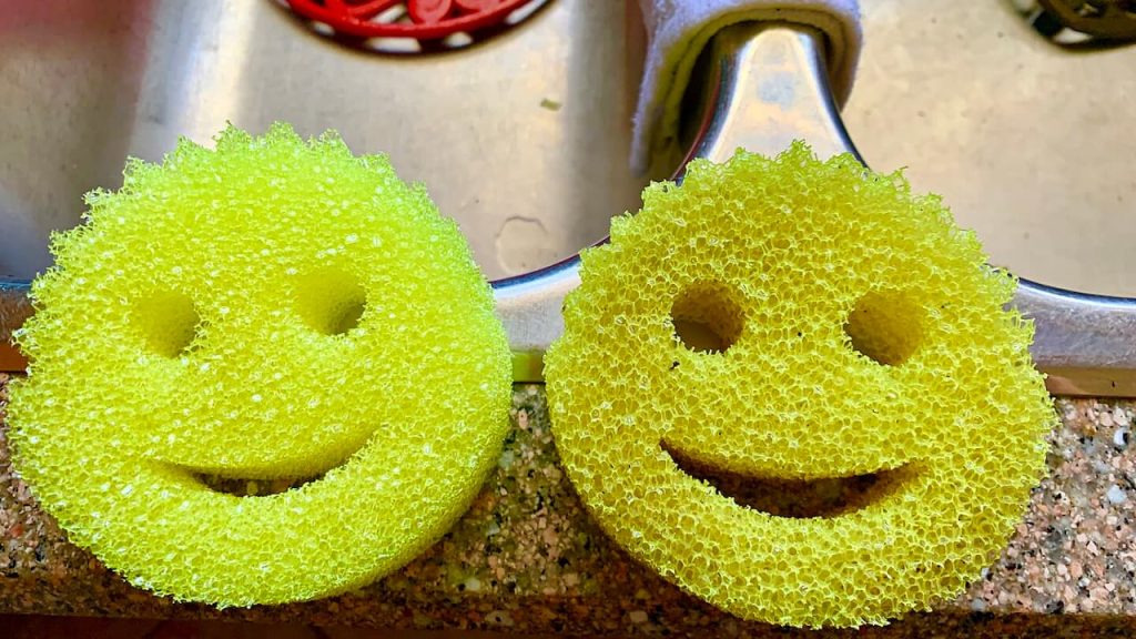Two yellow Scrub Daddy sponges with smiley face design sitting on granite countertop near kitchen sink faucet