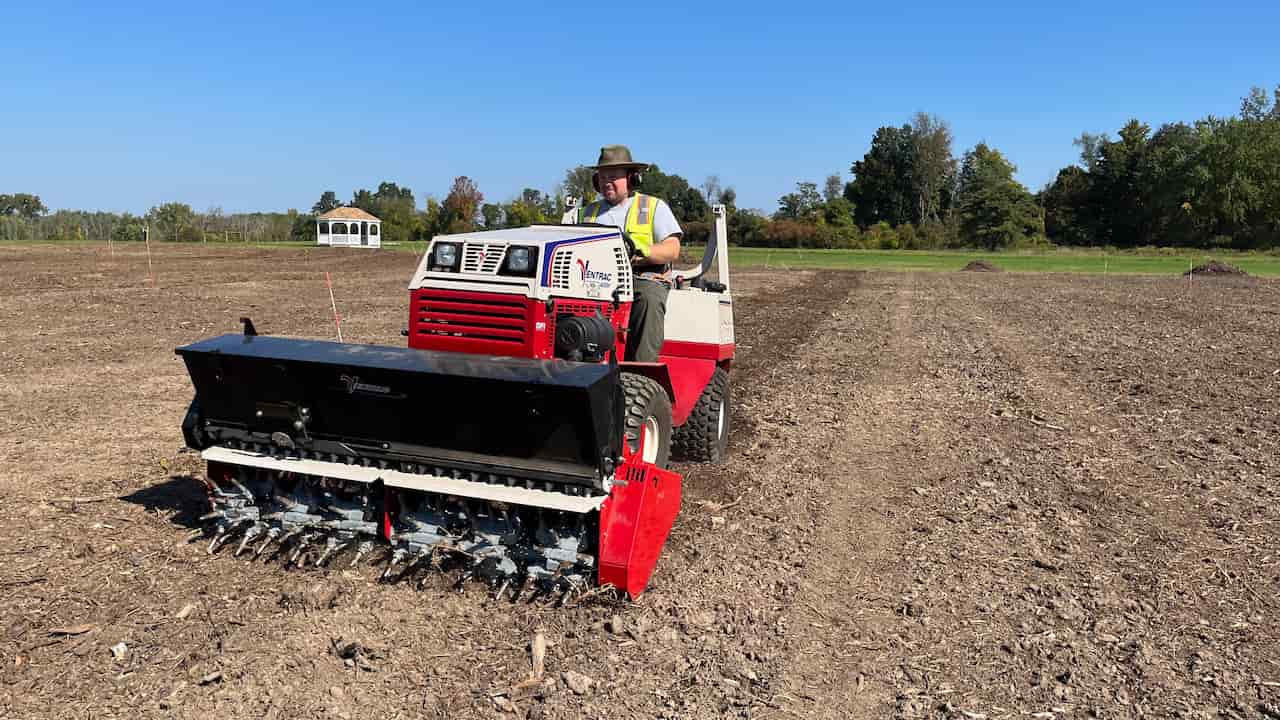 Man driving a Ventrac tractor across a dry farm field, using an aeration and seeding attachment, clear blue sky