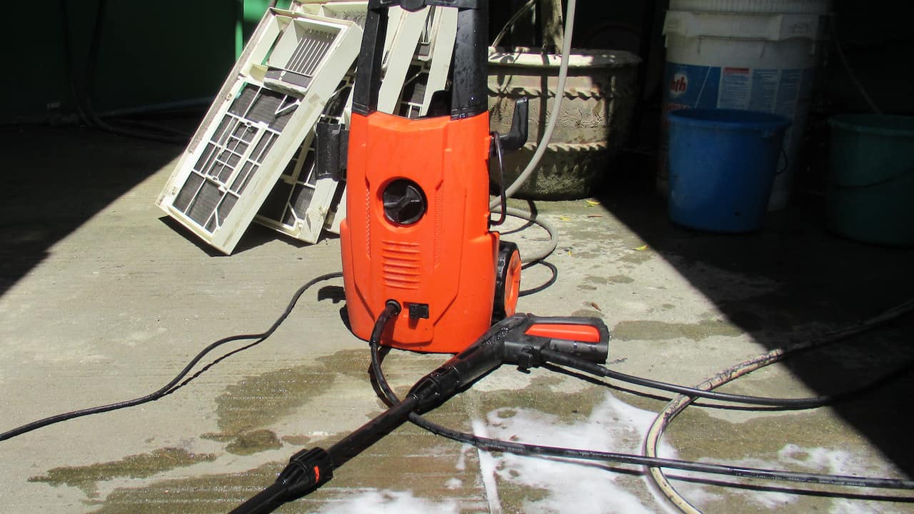 An orange and black electric pressure washer on a wet concrete floor, connected with hoses, surrounded by cleaning tools, buckets