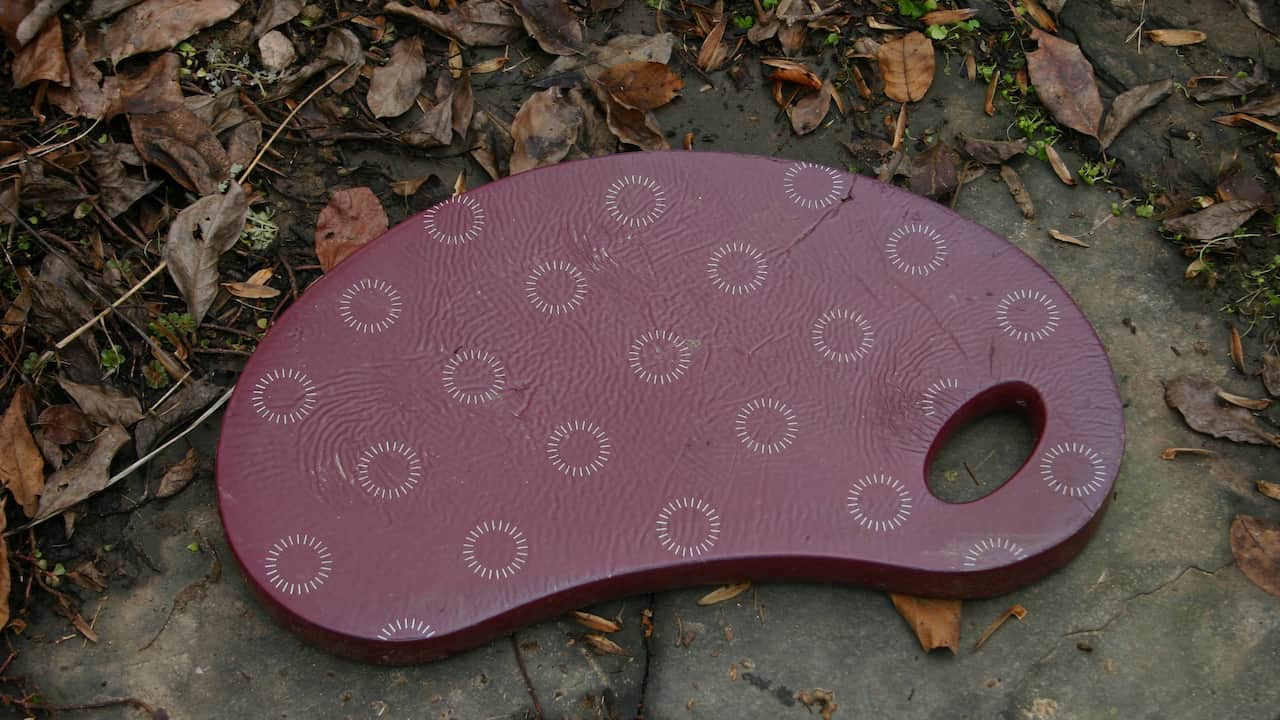 Maroon-colored foam kneeling pad with circular grip patterns and a built-in handle hole, placed on a stone surface surrounded by dry leaves