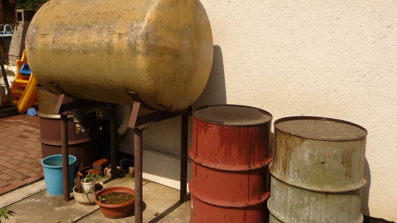 Rainwater collection system with a large horizontal tank on a metal stand, surrounded by three rusty metal barrels and small potted plants, placed against a beige wall