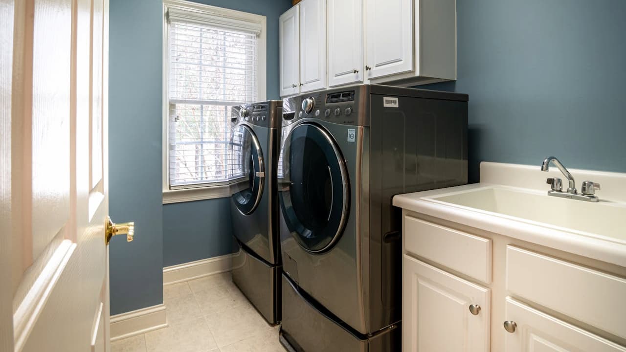 Blue laundry room with white cabinets, stainless steel front-loading washer and dryer, utility sink, window with blinds, tile flooring