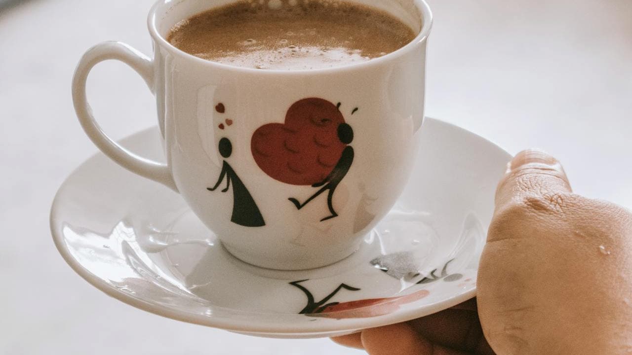 Close-up of a hand holding a white coffee cup and saucer, decorated with stick-figure art and a red heart design, filled with frothy hot coffee