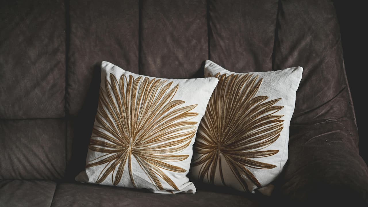 Two white throw pillows with brown palm leaf embroidery, resting on a dark brown cushioned couch