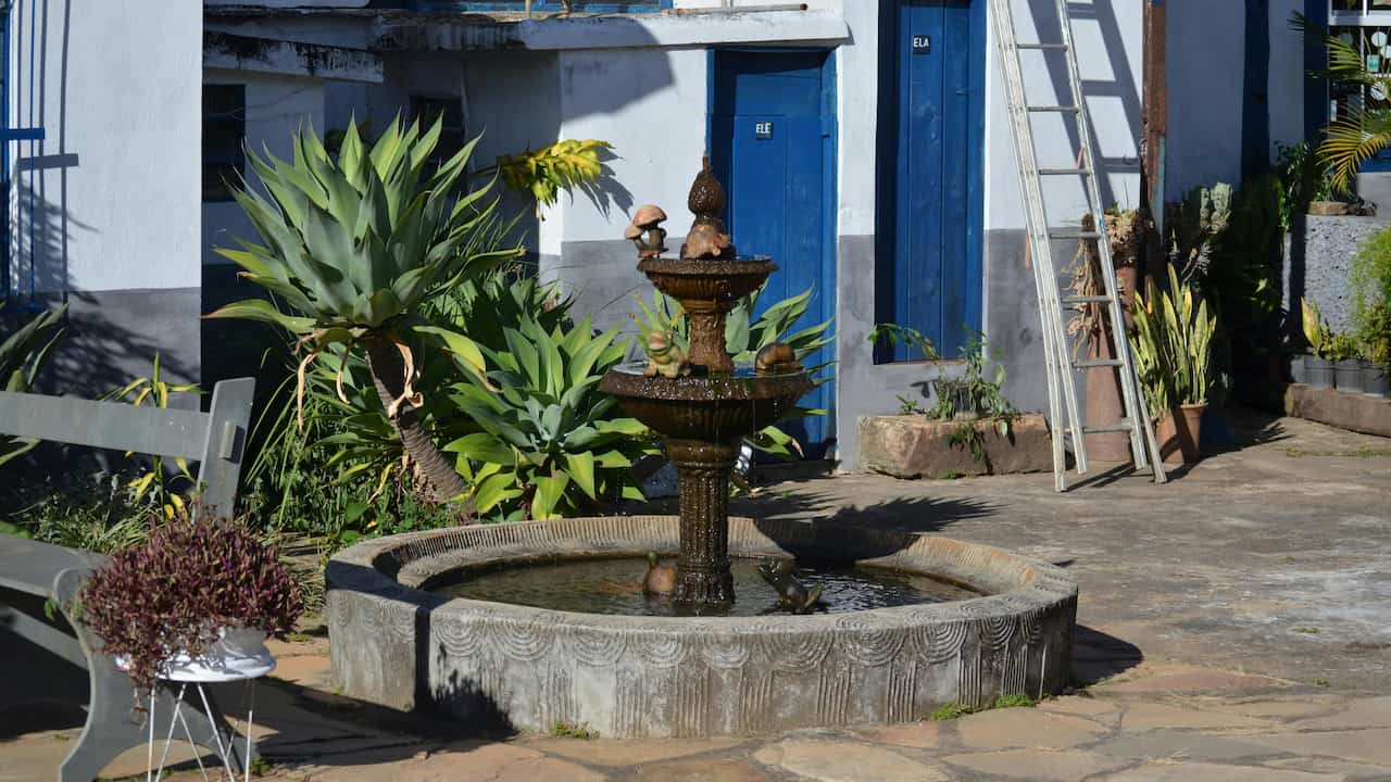Outdoor stone water fountain in a courtyard, surrounded by lush green plants, a bench, a ladder, and blue restroom doors labeled "ELE" and "ELA" in the background