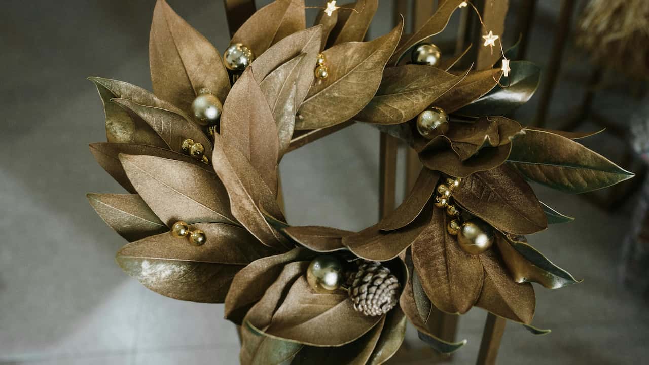 Brown magnolia leaf wreath decorated with gold ornaments and a pine cone, displayed indoors on a wooden stand