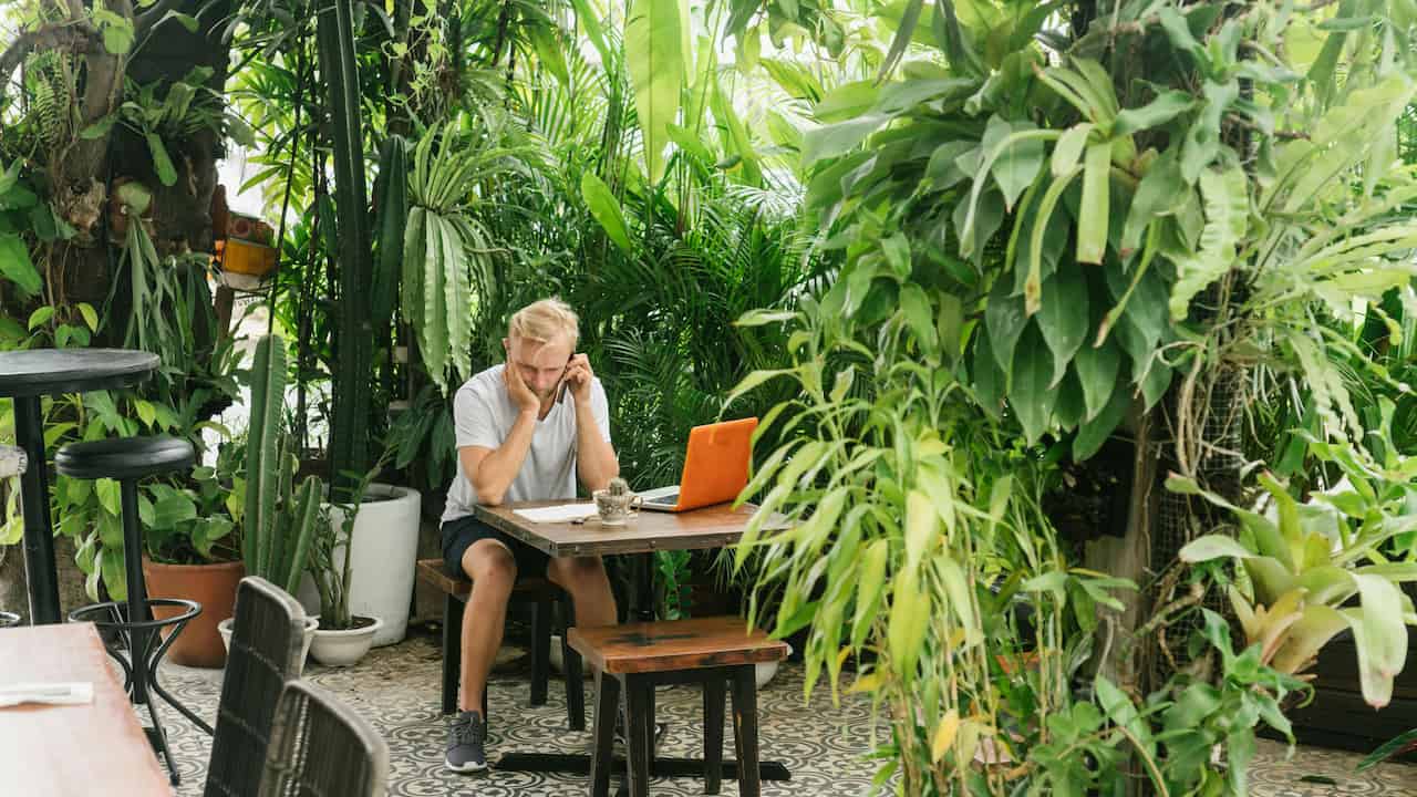 Man sitting at a wooden table surrounded by lush green tropical plants, using a phone while looking at a laptop with an orange cover