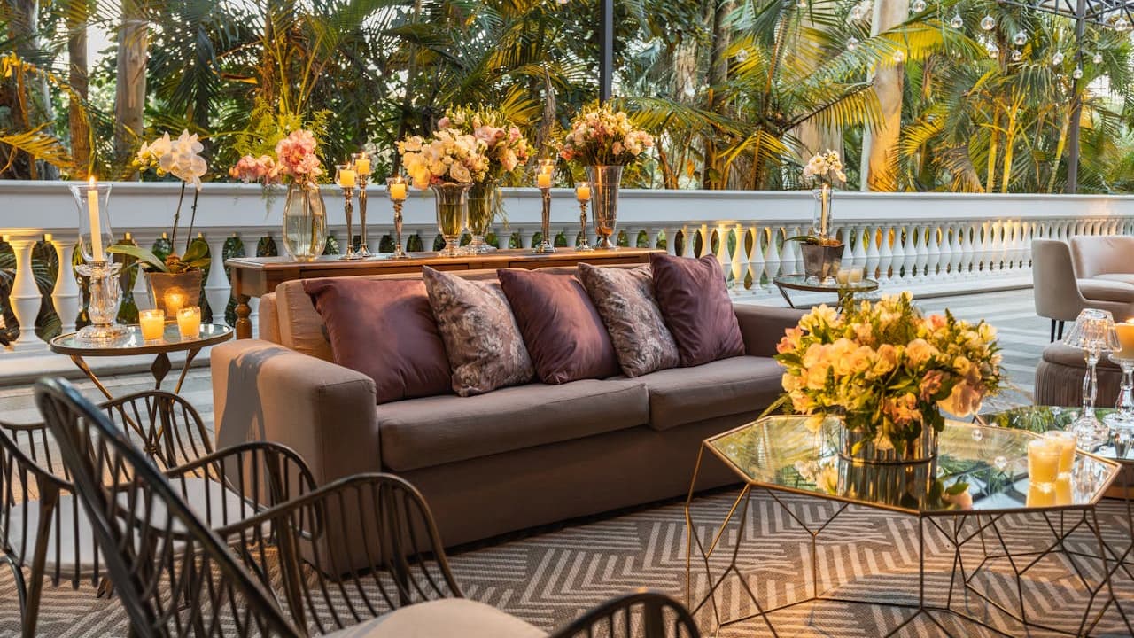 Elegant outdoor terrace lounge with gray sofa, floral arrangements, candles, and tropical palm trees in golden sunset lighting
