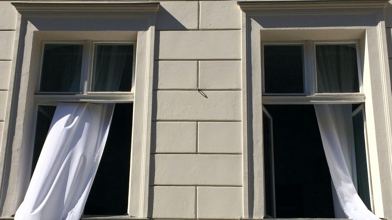 Two open windows with white curtains billowing outward, mounted on a light-colored building facade, lit by sunlight