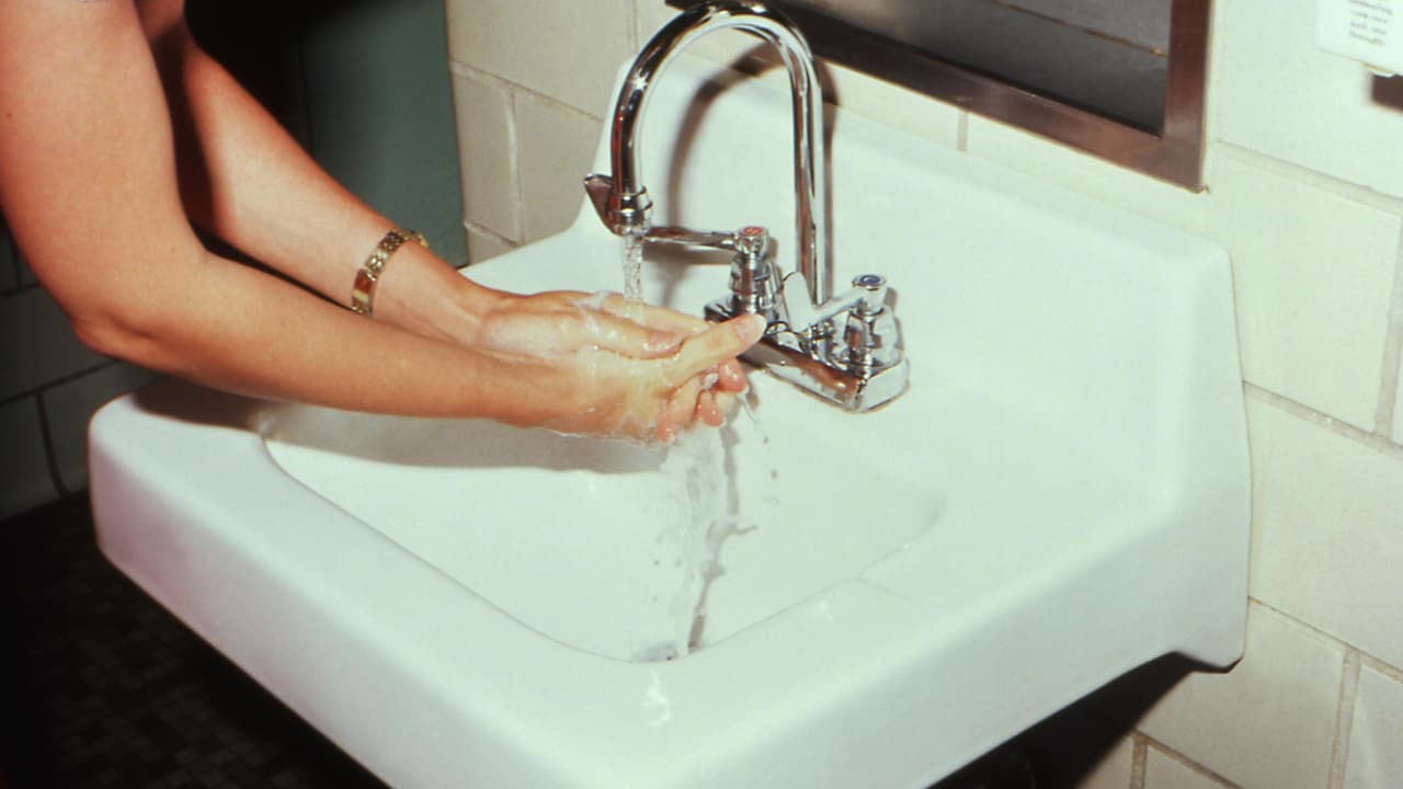 Person washing hands under running water at a white ceramic sink in a tiled restroom