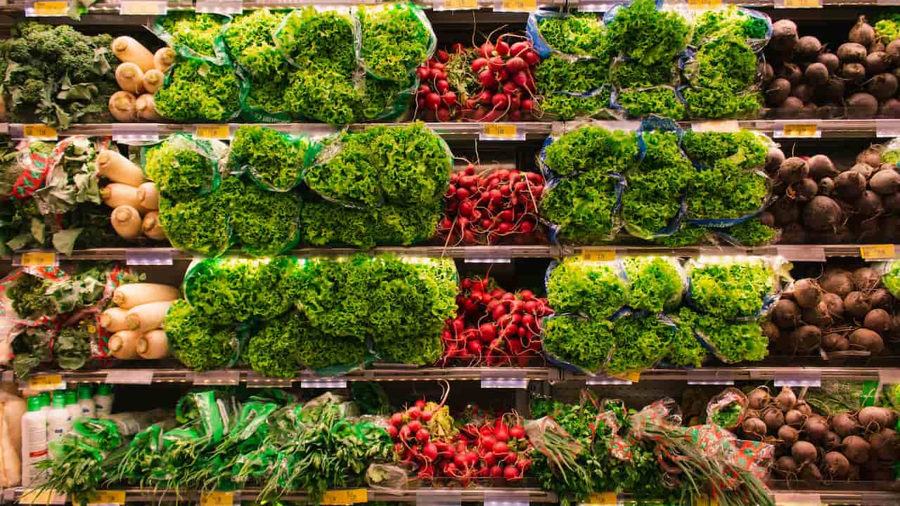 Supermarket produce section, neatly arranged vegetables on shelves, including lettuce in plastic wrap, radishes, daikon, kale, beets, and herbs, brightly lit display