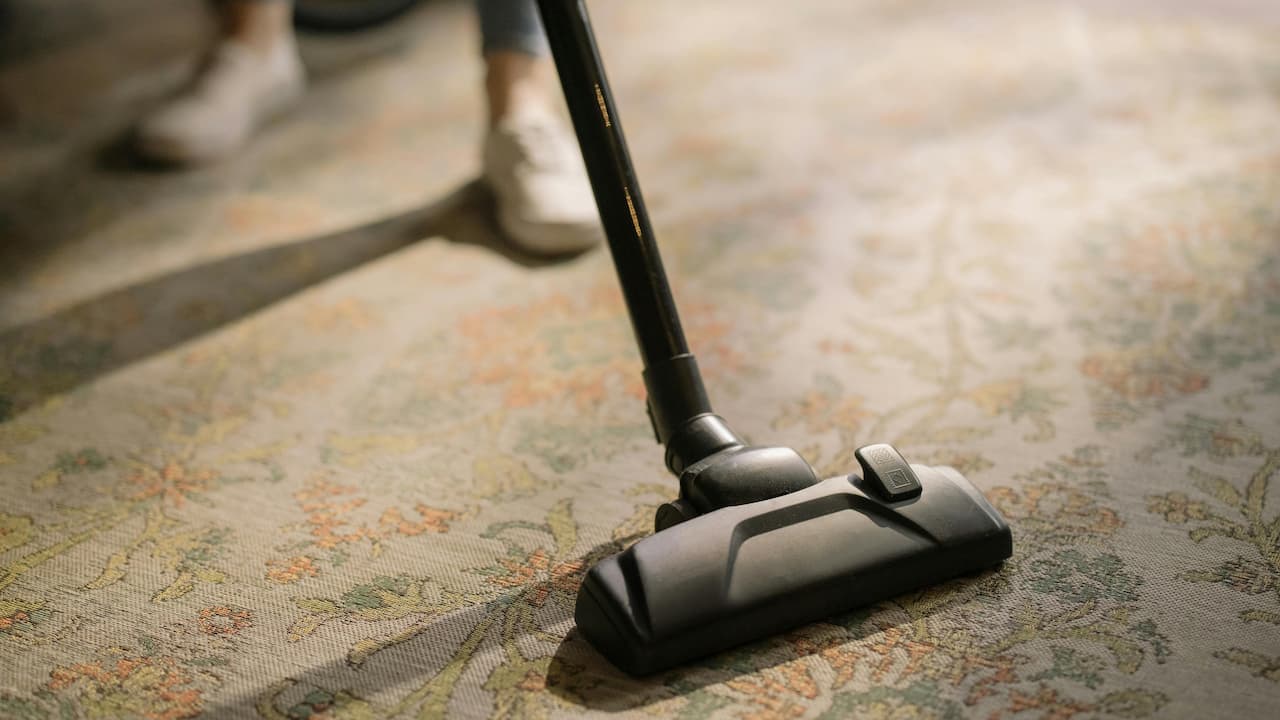 Person vacuuming a patterned carpet with a black vacuum cleaner, wearing white shoes and jeans, soft lighting from the side