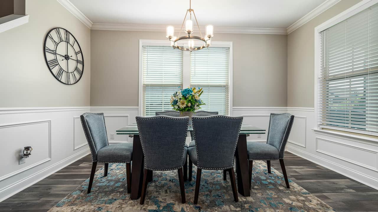 Elegant dining room with six gray upholstered chairs, a glass-top table, floral centerpiece, large wall clock, chandelier, and white-trimmed windows with blinds