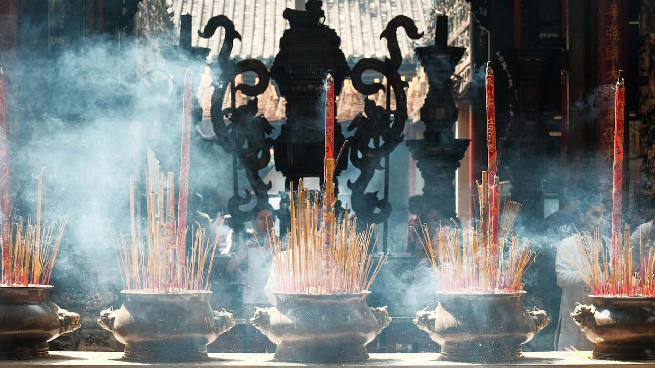 Bronze incense burners filled with lit incense sticks, with curling smoke rising in a traditional temple setting