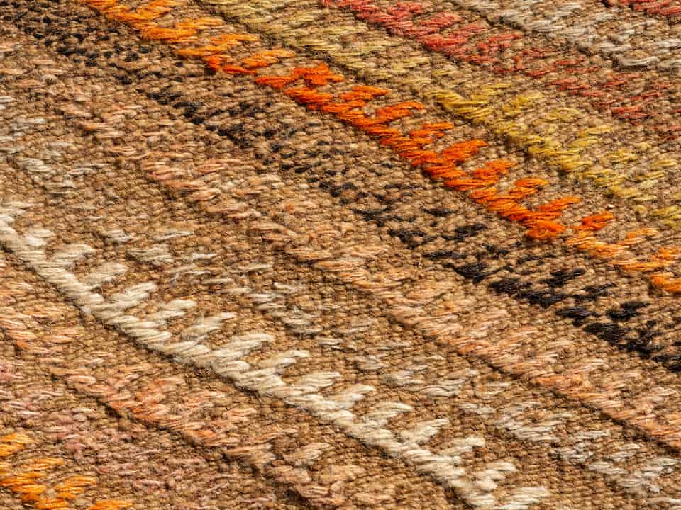 Close-up of a woven textile with earthy tones, featuring diagonal patterns in brown, beige, orange, yellow, and cream