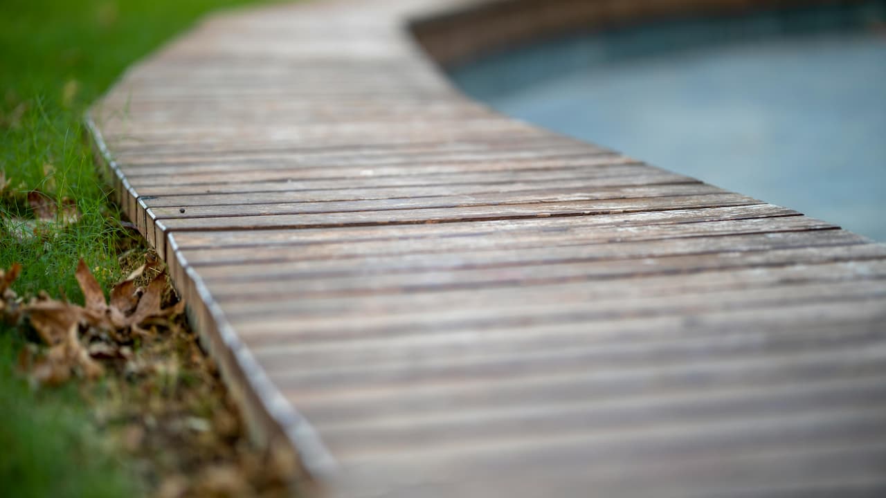 Curved garden pathway made of wooden planks, bordered by green grass on one side and a body of water on the other