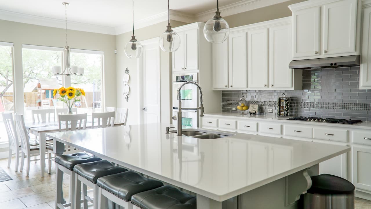 Bright modern kitchen with white cabinets, a large white island with black barstools, stainless steel appliances, and pendant lighting