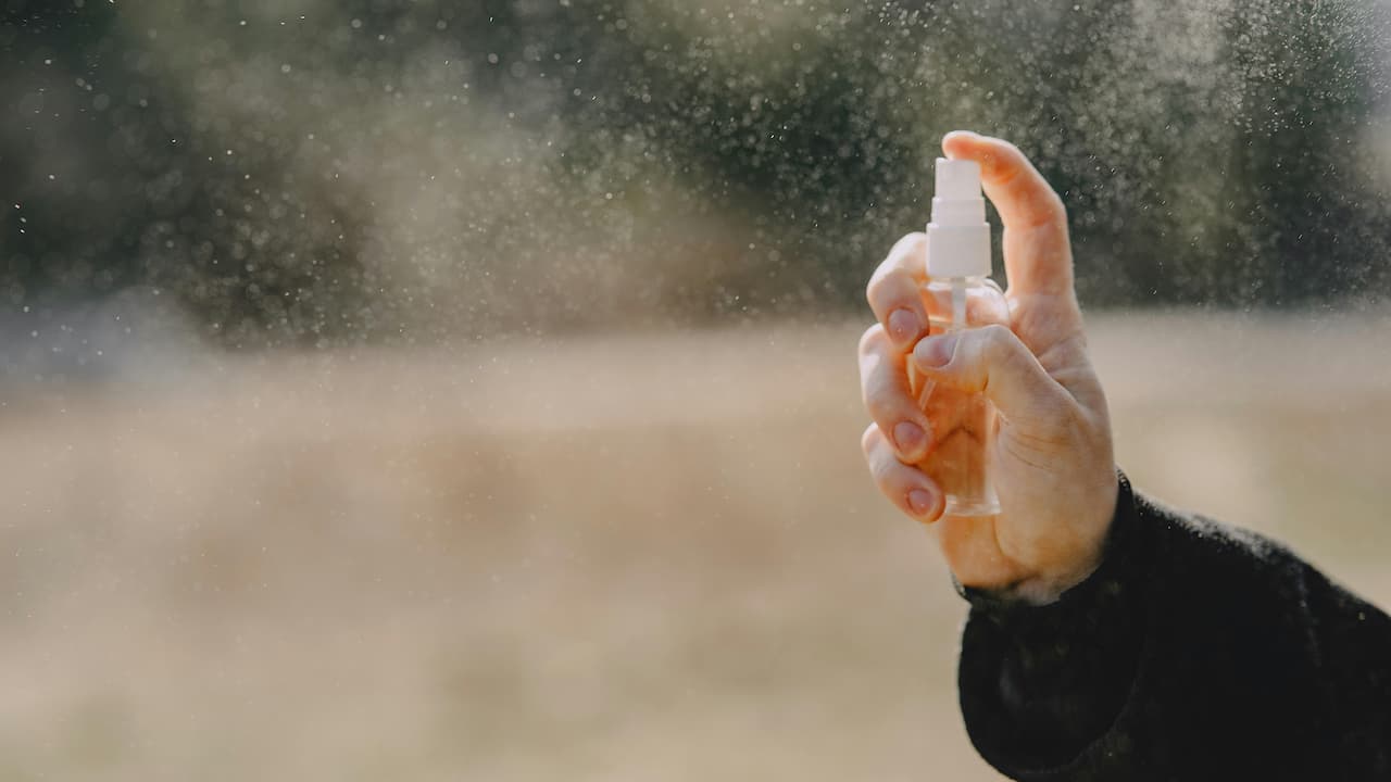 Hand spraying a clear mist from a small transparent plastic bottle, mist visible in the sunlight against a blurred outdoor background