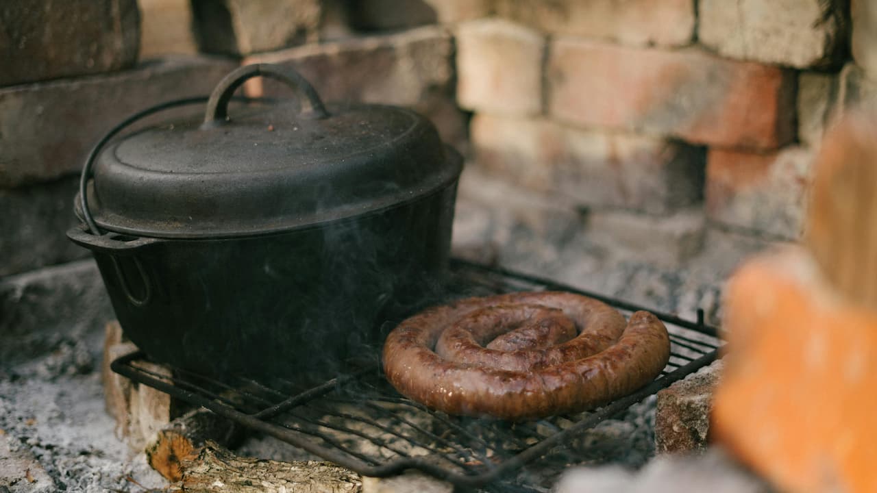 Coiled sausage and cast iron pot cooking on a rustic brick barbecue with visible smoke rising from the grill