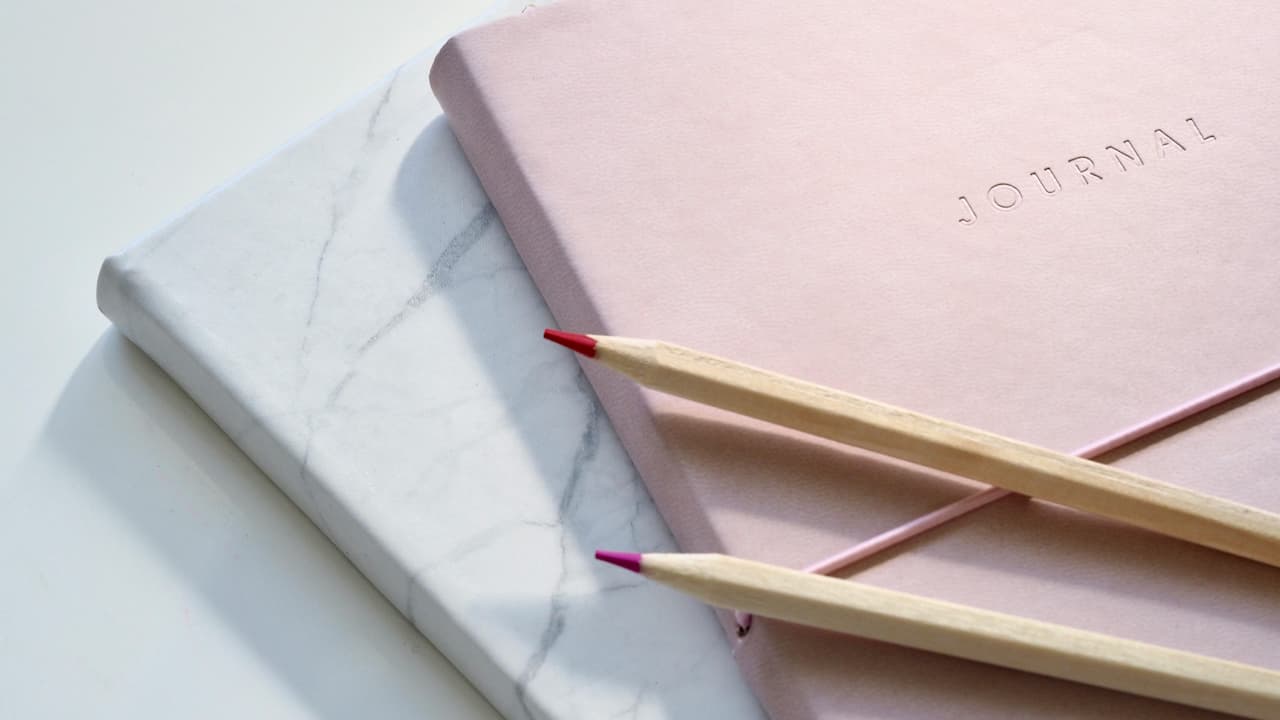 Pink journal and marble-patterned notebook, two colored pencils, soft lighting, clean white background