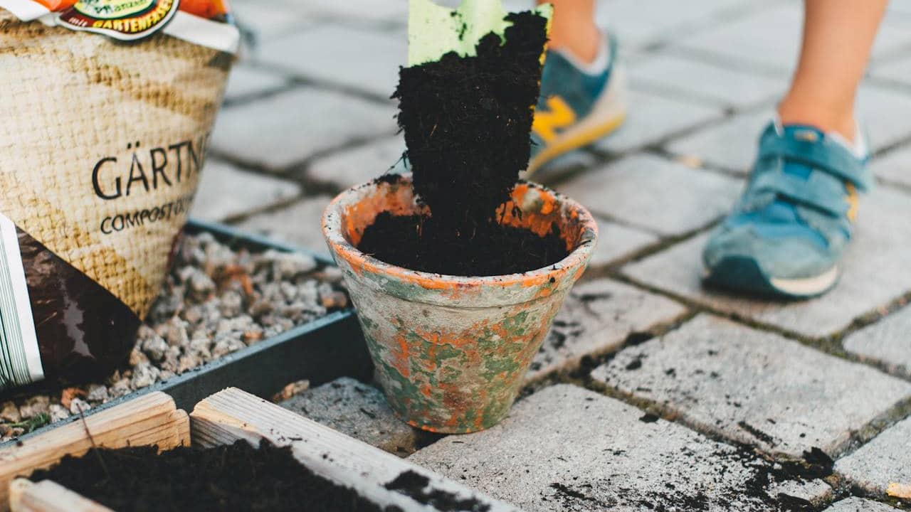 A green garden trowel pours rich black soil into a rustic, weathered terracotta pot, surrounded by compost bags and a wooden pallet-style planter on a stone-paved surface