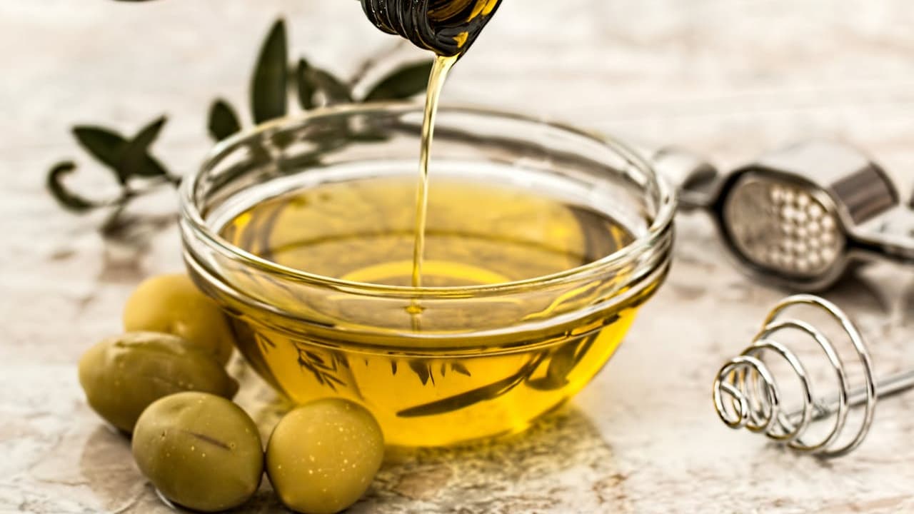 Olive oil being poured into glass bowl surrounded by green olives, garlic press, and olive branch decoration