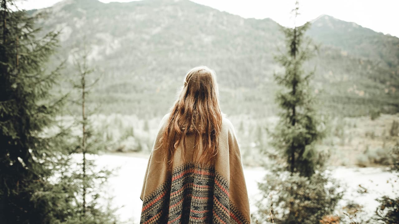 Woman with long brown hair wearing a patterned blanket, standing outdoors and facing a mountain landscape with evergreen trees