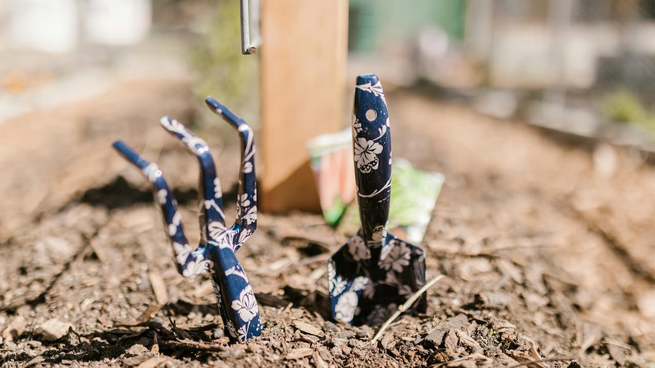 Two floral-patterned gardening tools—a hand rake and a trowel—are planted upright in soil mulch in a raised garden bed, with seed packets and a wooden stake blurred in the background
