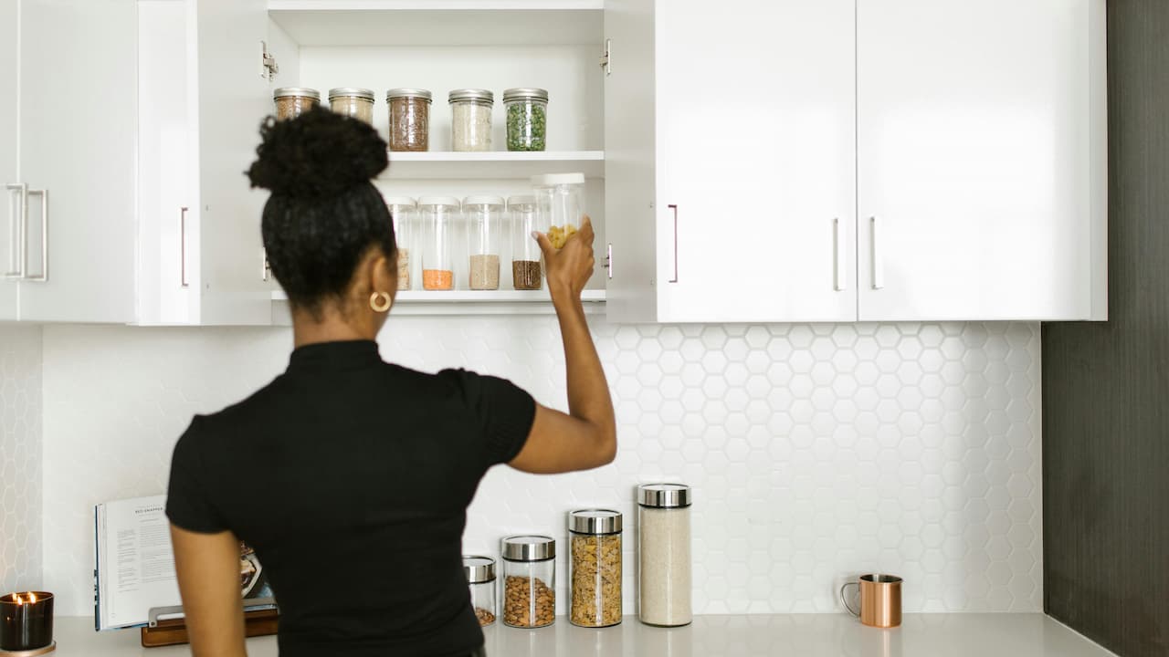 Woman organizing food jars in a white kitchen cabinet with glass containers of grains and legumes
