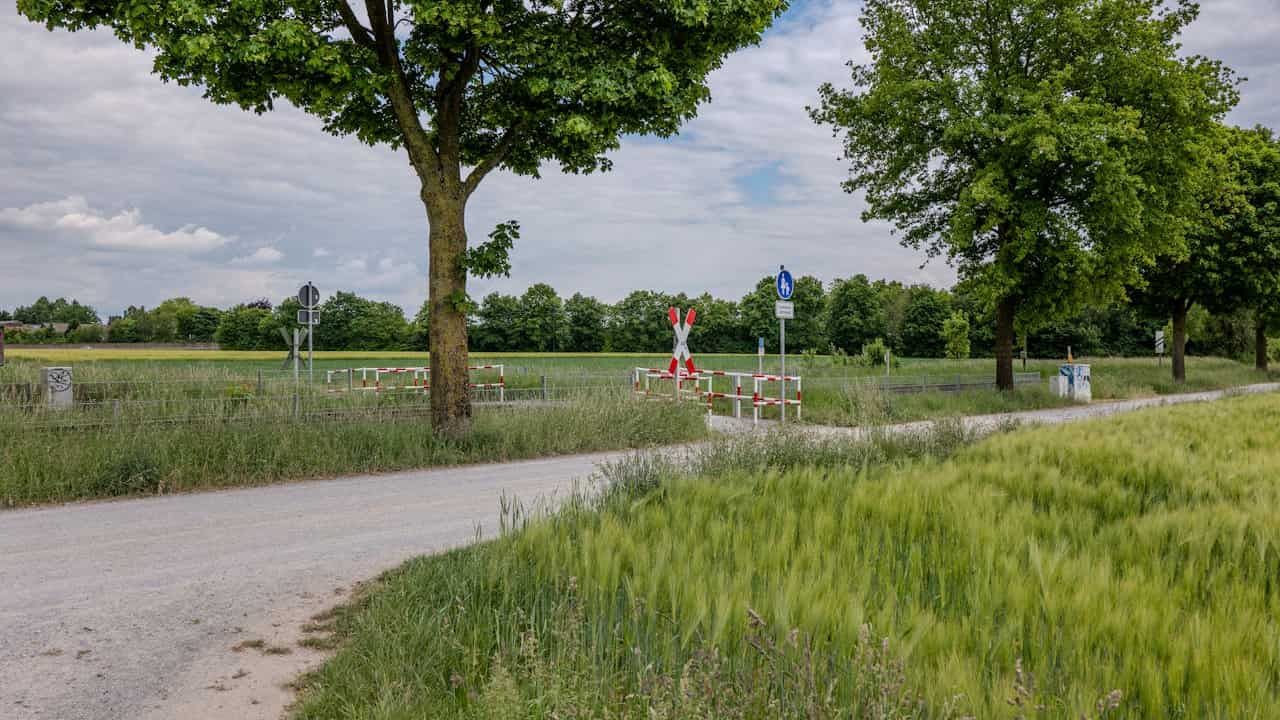 Gravel road with a rural railroad crossing, bordered by green fields, tall grass, and trees under a cloudy sky, with warning signs and red-and-white safety barriers