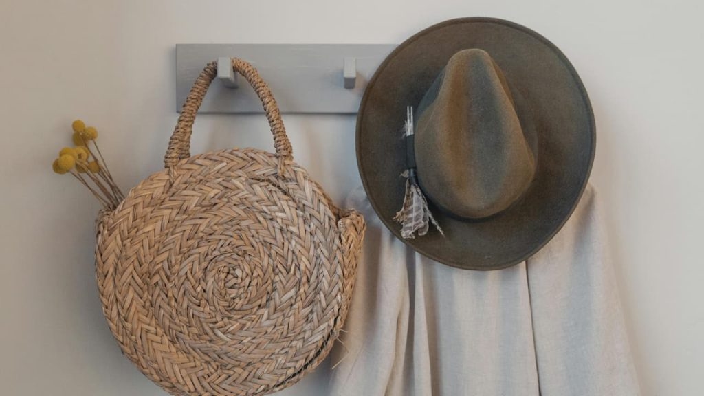 woven round bag with yellow flowers, felt hat with tassel, and light scarf hanging on wall hooks
