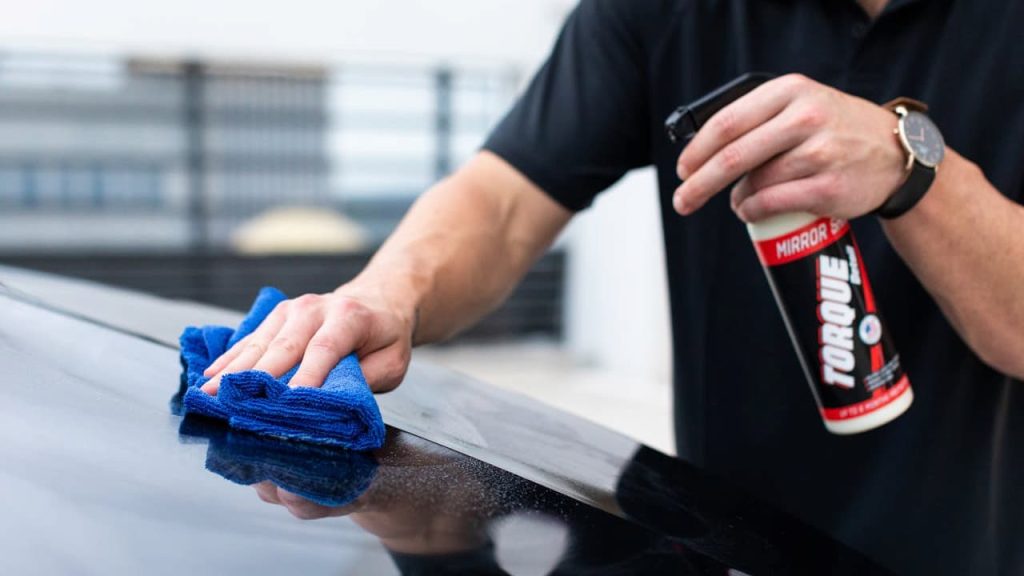 Person cleaning car surface with blue microfiber cloth while holding spray bottle of automotive cleaning product