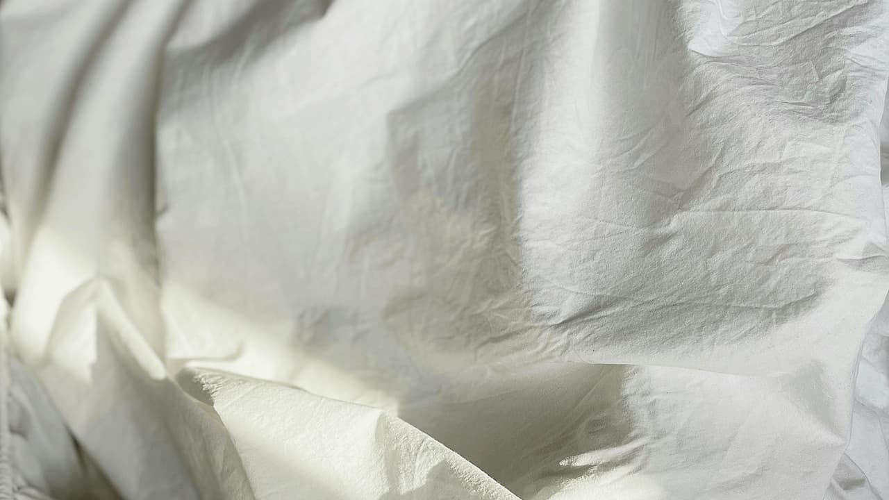 Wrinkled white bed sheets, softly lit by natural sunlight, creating gentle shadows and folds