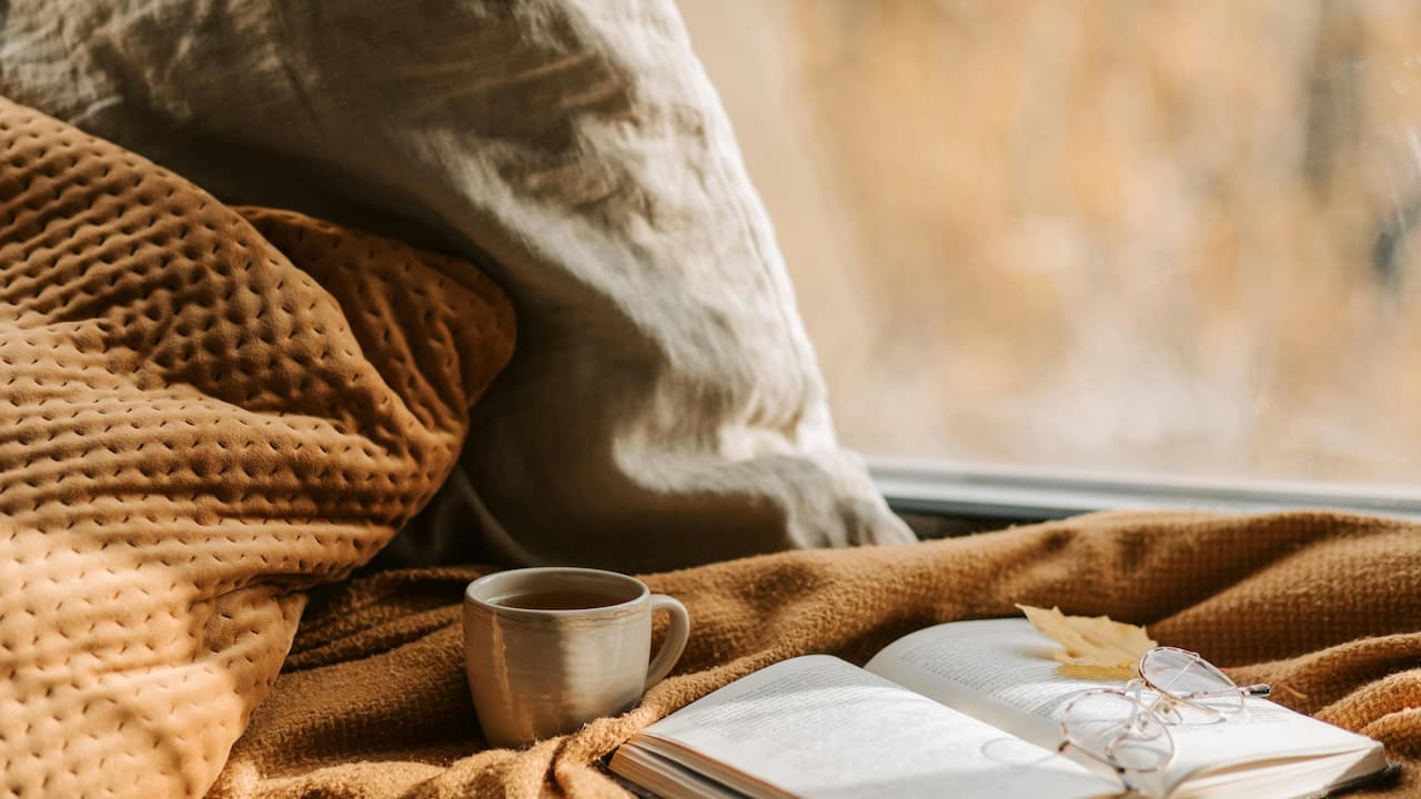 Cozy reading nook with a warm brown blanket, soft pillows, a cup of tea, an open book with reading glasses resting on it