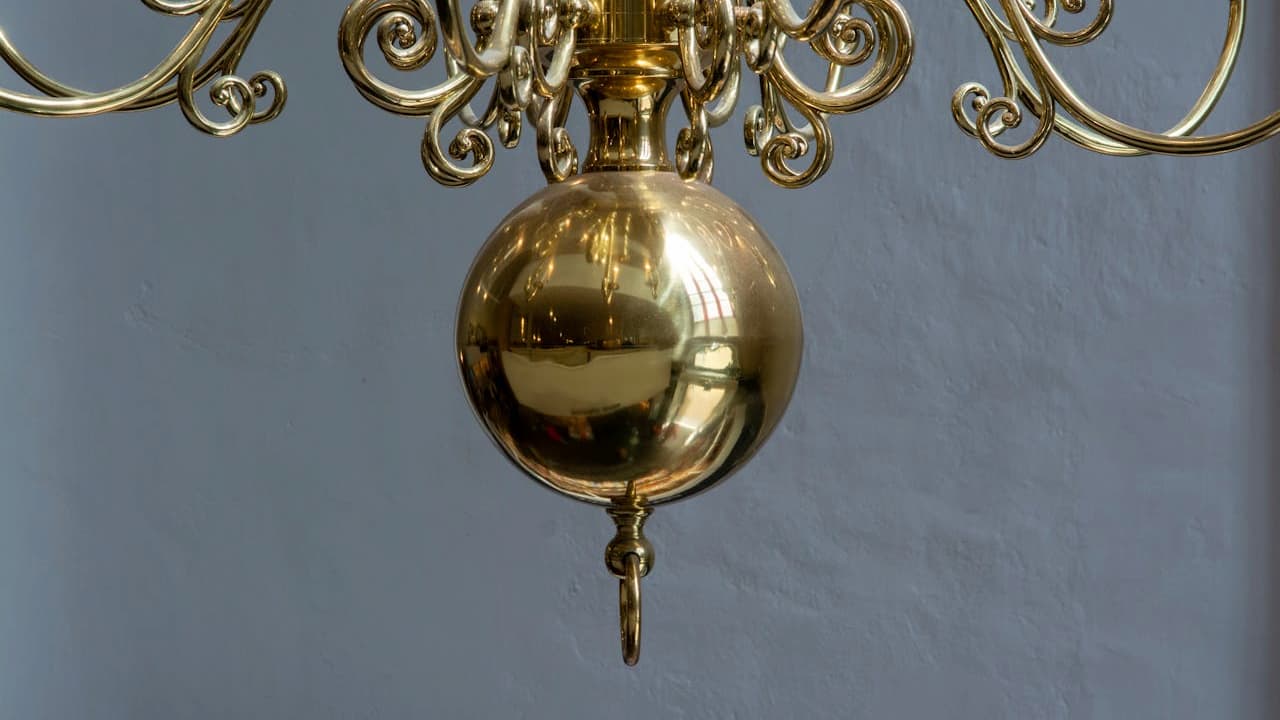 Ornate brass chandelier with decorative scrollwork arms and polished spherical pendulum hanging against gray wall