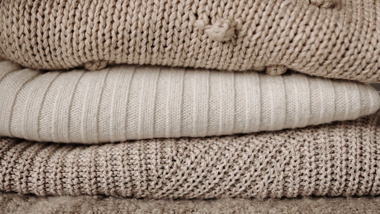 Stack of folded knit blankets in neutral beige and cream tones, featuring varied textures including ribbed, waffle, and popcorn knit patterns