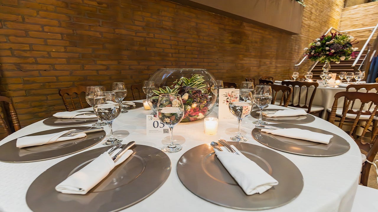 Round table set for a formal event, white tablecloth, silver chargers, white napkins neatly rolled with silver napkin rings, wine glasses and cutlery arranged uniformly, glass bowl centerpiece with flowers and candle, brick wall background, warmly lit indoor setting