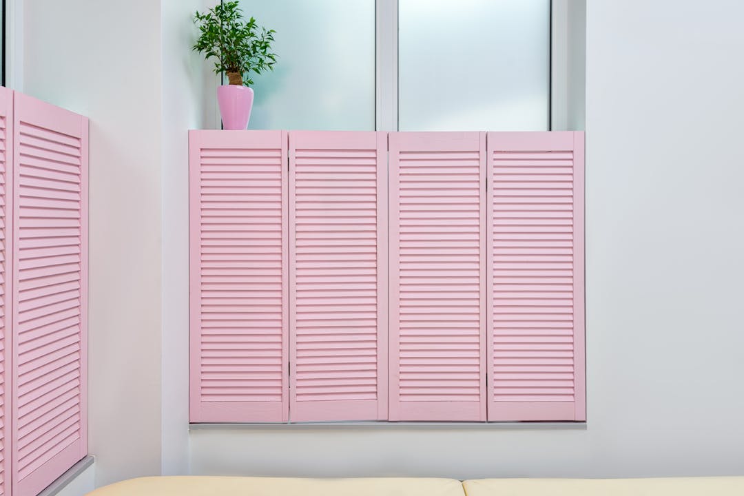 Pink louvered cabinet doors mounted on white wall, small potted plant on top, partial view of matching pink shutters on left