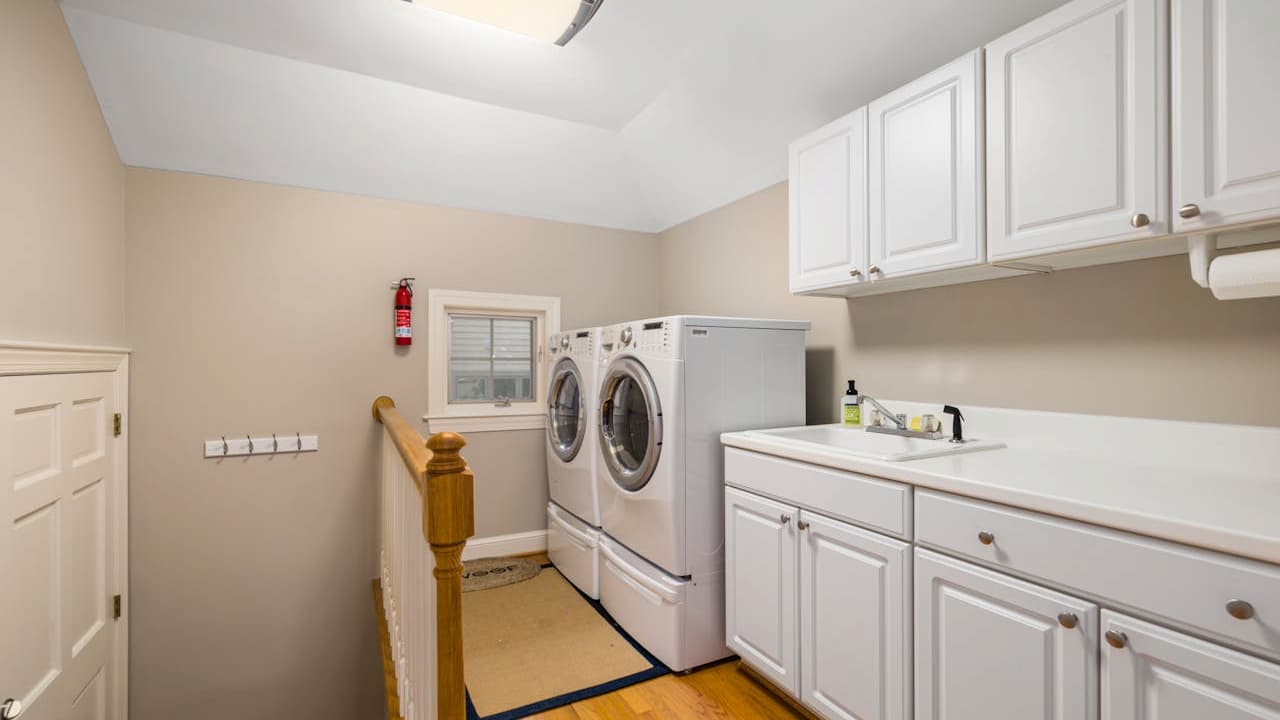 White laundry room with front-loading washer and dryer, utility sink, white cabinets, wooden stair railing visible, fire extinguisher on wall