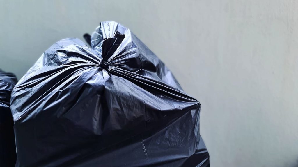 Black plastic garbage bags tied with drawstrings placed against light gray wall, ready for waste collection or disposal