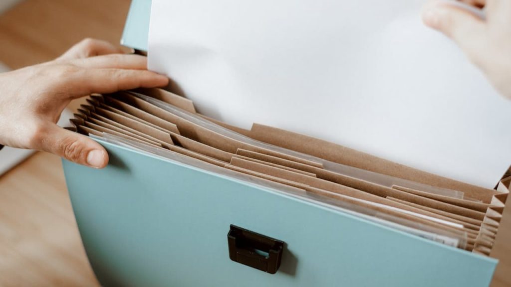 Person's hands organizing manila file folders inside light blue filing box with black metal label holder on front