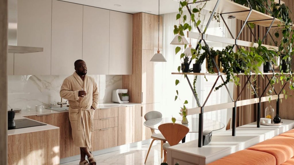 Man in beige bathrobe holding cup in modern kitchen with wood cabinets, white countertops, and tiered plant stand for layering
