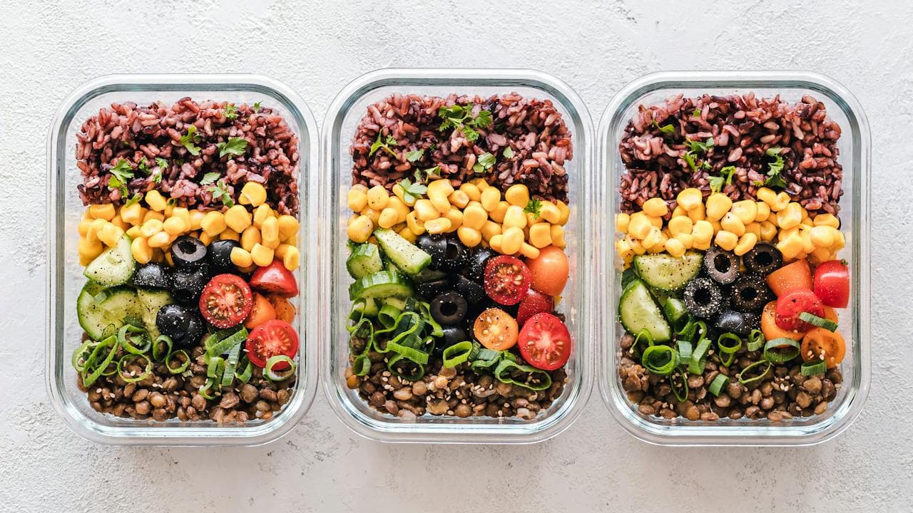 Three glass meal prep containers filled with layered vegetarian bowls containing red rice, chickpeas, vegetables, and fresh herbs